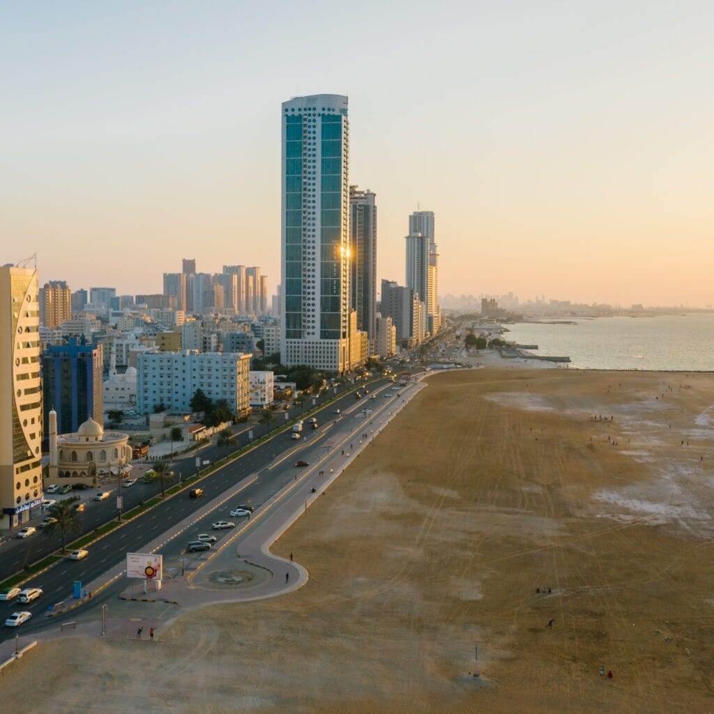 Low Cost Business Setup in Dubai A coastal cityscape at sunset in Dubai Offshore, featuring a wide road lined with modern high-rise buildings on one side and a vast sandy area on the other, extending toward the sea. The tall structures reflect the golden light of the setting sun, highlighting the region's growth and opportunities often supported by leading business setup consultants in Dubai Offshore
